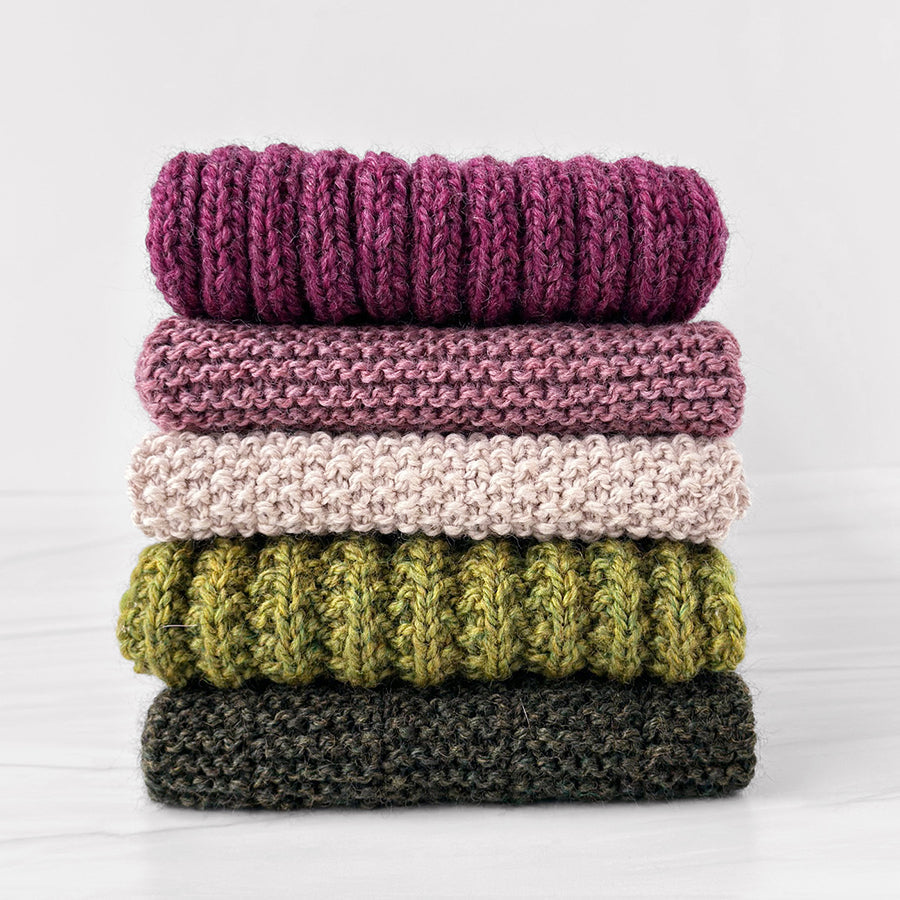 Five folded scarves knitted in Blue Sky Fibers Woolstok, stacked to show ribbed, garter, and seed stitch textures in purple, lilac, beige, and green.