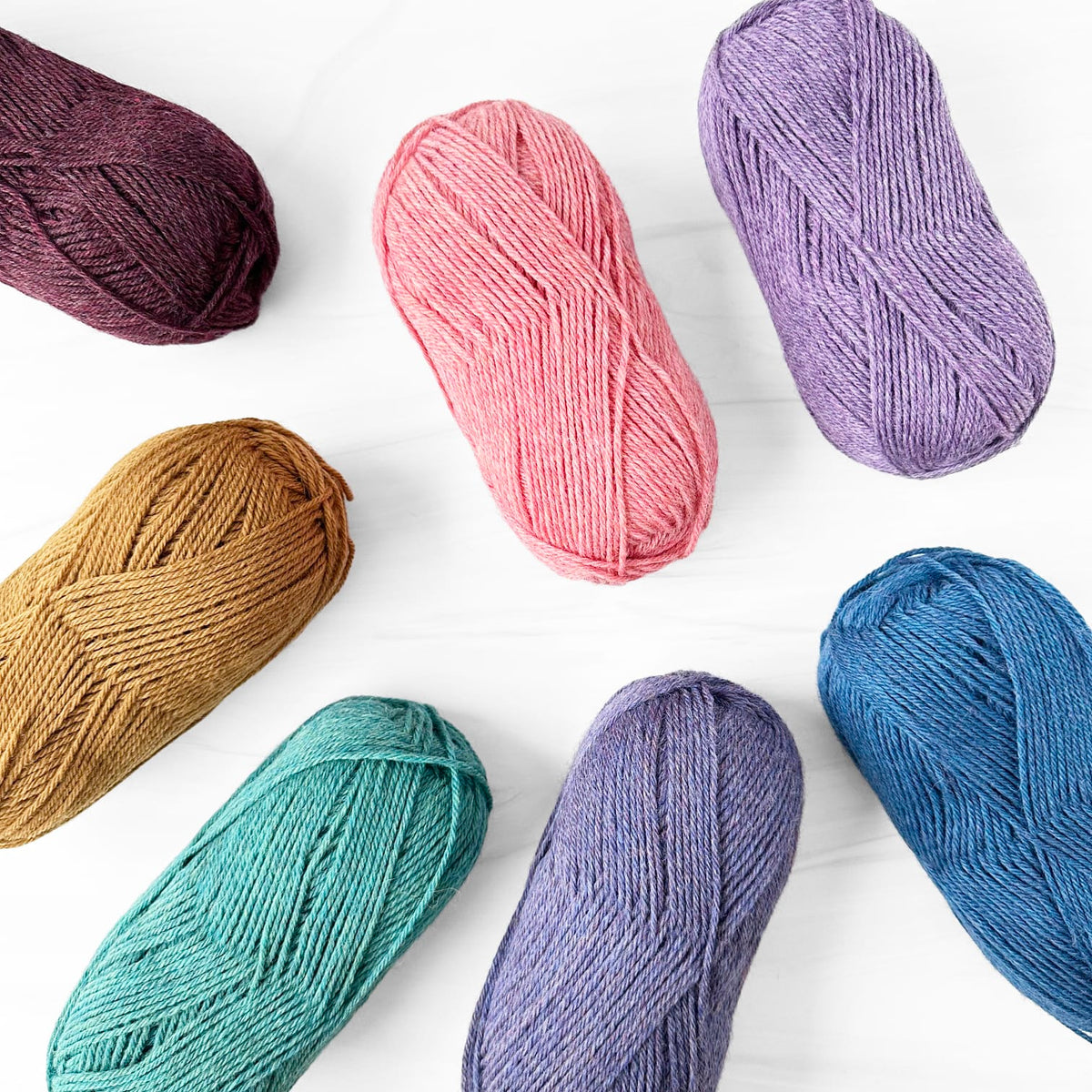 Six skeins of Berroco Lanas yarn in assorted colors arranged on a white background: burgundy, pink, lavender, tan, teal, and medium blue.
