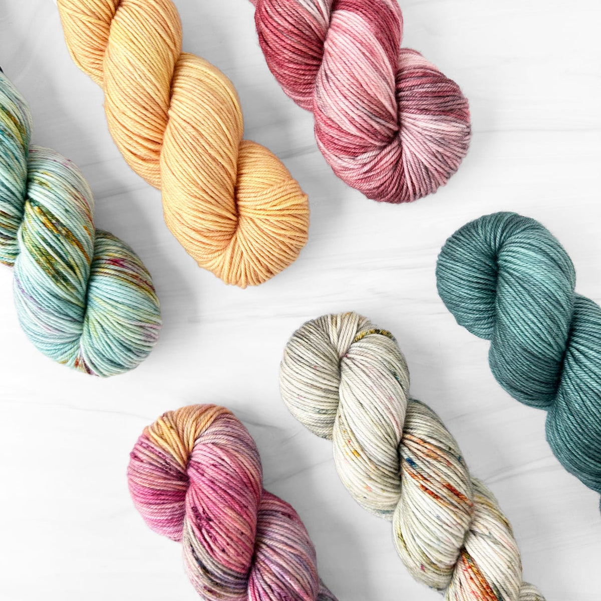 Six hanks of Fiberstory Core DK variegated yarn arranged on a white wood background. The yarns come in multiple colors including pastel blue-green, soft yellow, dusty rose, cream with orange speckles, and teal with pink and brown variegation.