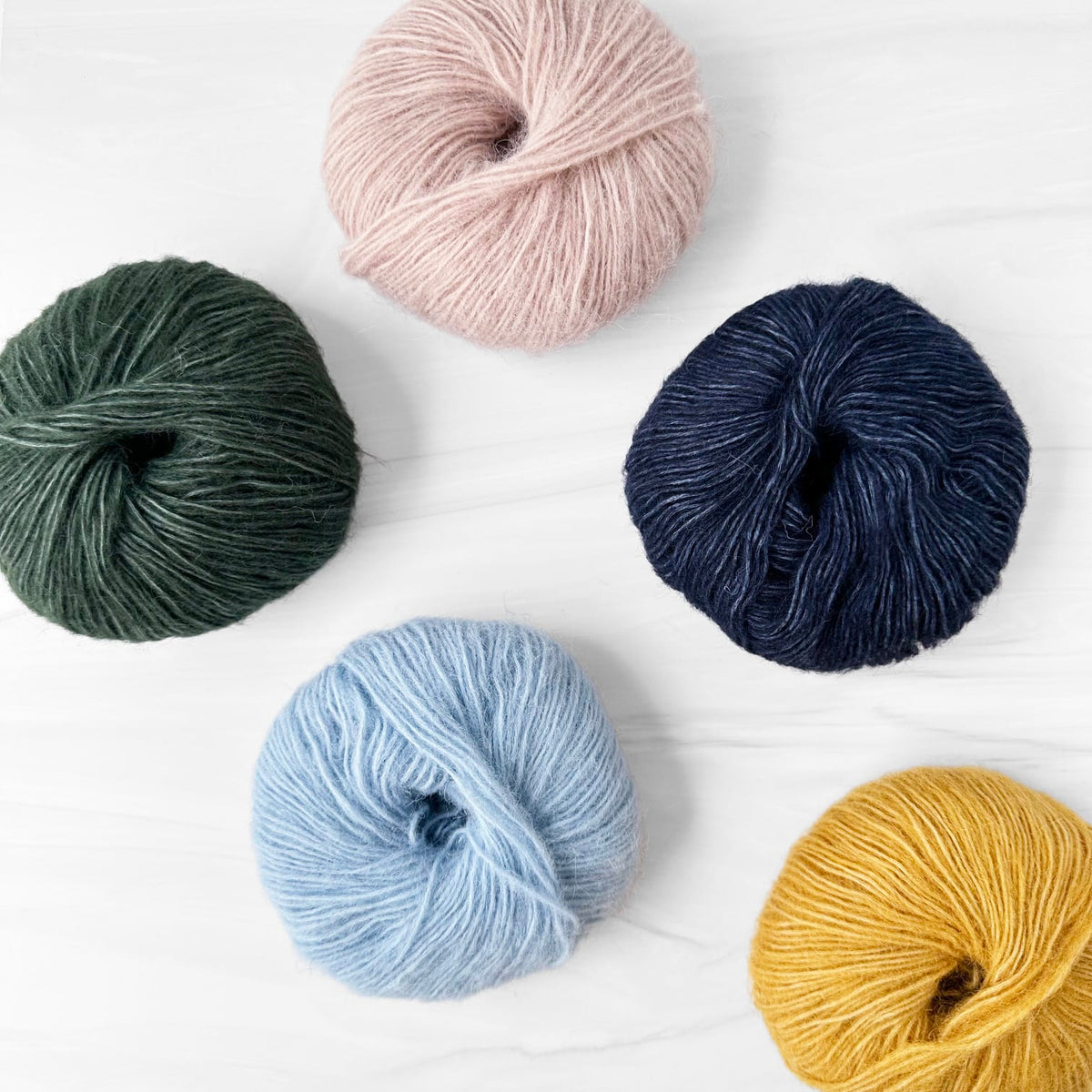 Five skeins of Alpaca Cotton yarn in different colors arranged on a white wood background: dark green, light pink, navy blue, light blue, and mustard yellow.