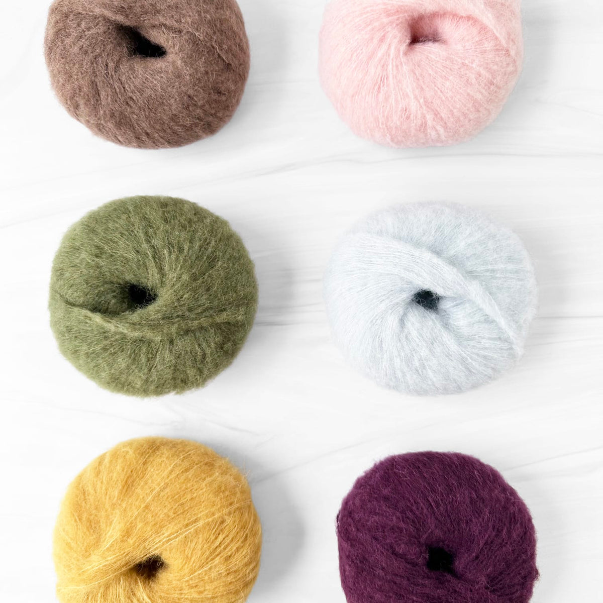 Six balls of Alpaca Haze yarn arranged in a 2x3 grid on a white wood background. The yarn colors are brown, light pink, sage green, pale blue, mustard yellow, and deep purple.