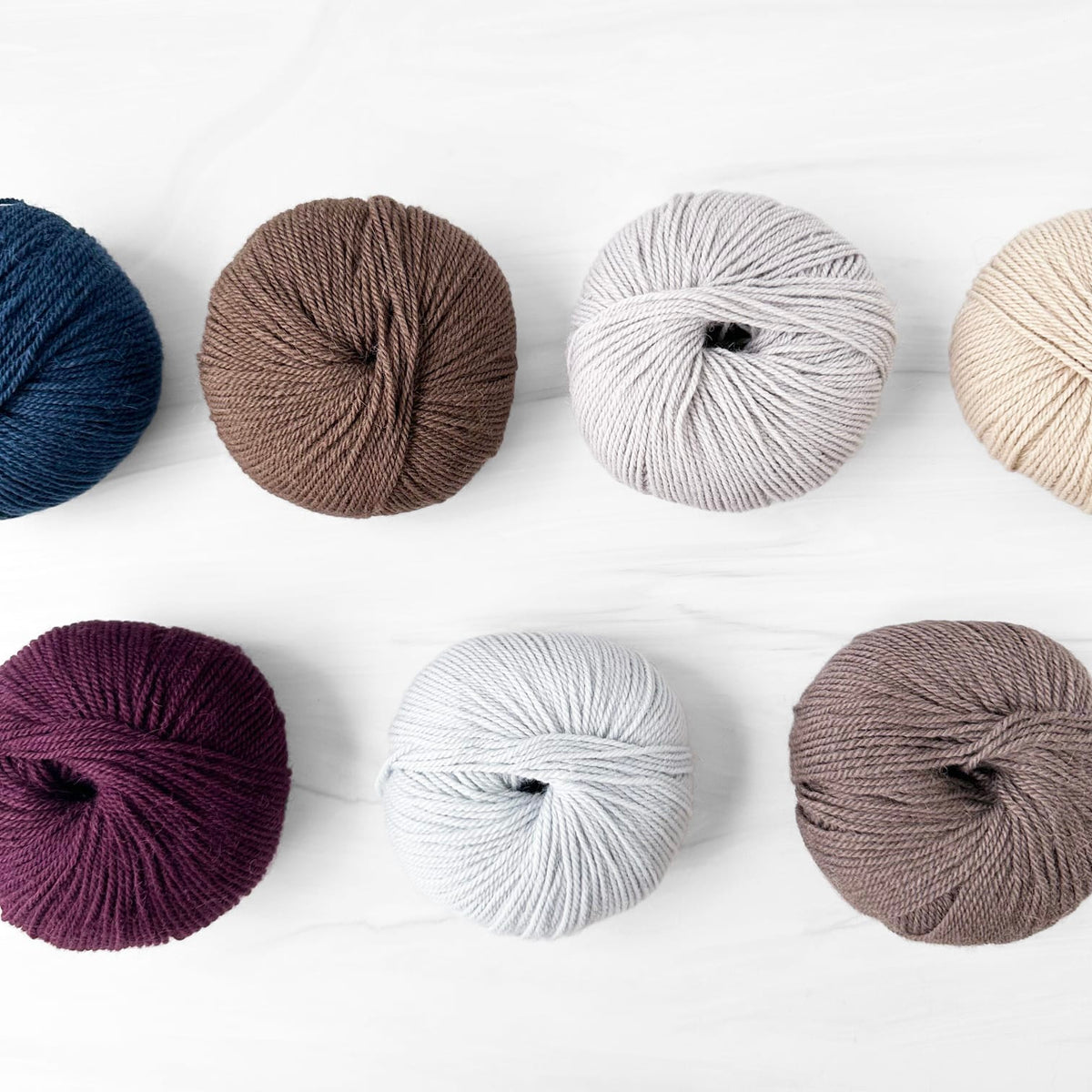 Six skeins of Alpaca Wool yarn arranged in two rows on a white marble surface, shown in navy blue, brown, light gray, cream, deep purple, pale blue, and taupe.