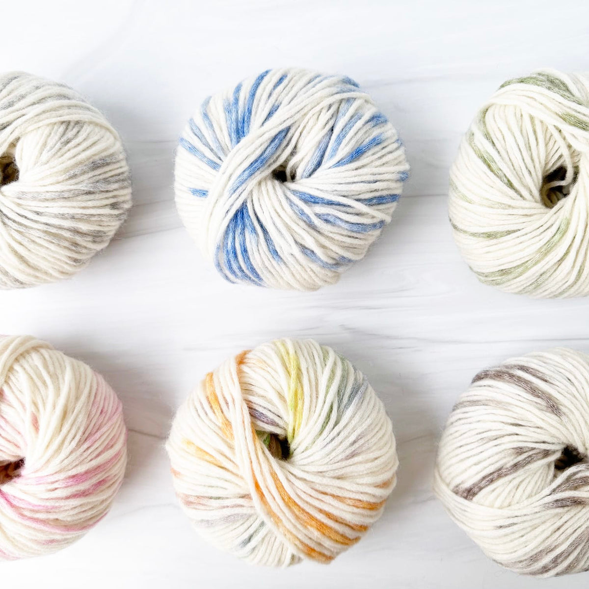 Six skeins of MODE at Rowan Colormix yarn arranged in two rows on a white wood background. The yarns feature variegated colors including blue, pink, yellow, green, and gray on an off-white base.