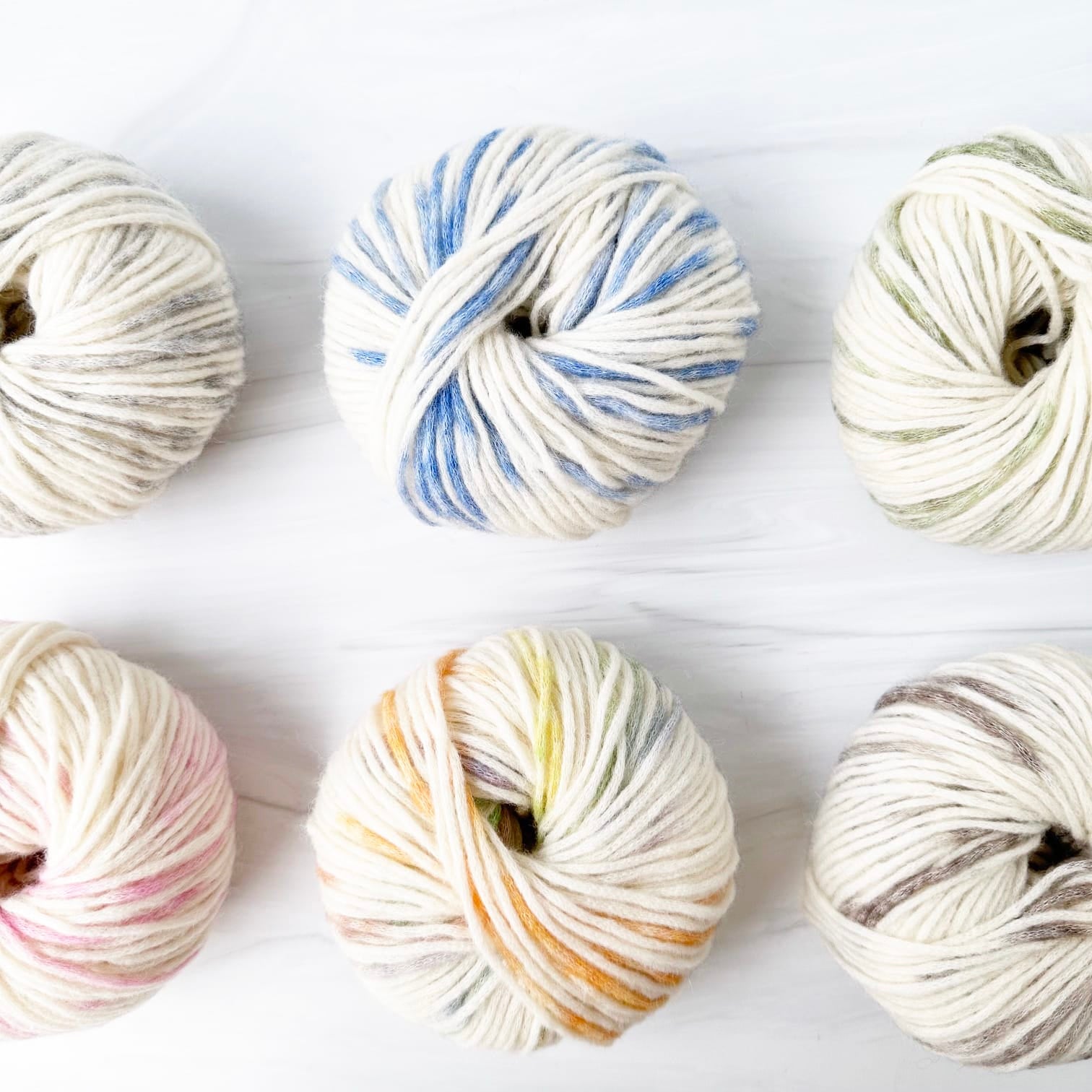 Six skeins of MODE at Rowan Colormix yarn arranged in two rows on a white wood background. The yarns feature variegated colors including blue, pink, yellow, green, and gray on an off-white base.