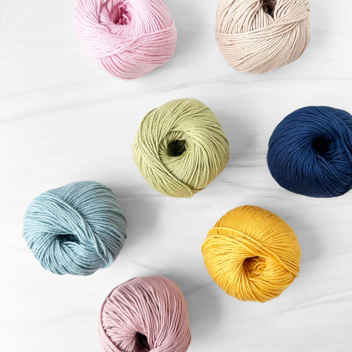 Six balls of MODE at Rowan Cotton DK yarn in assorted pastel and neutral colors are arranged on a light white marble surface. The colors include light pink, cream, sage green, navy blue, light blue, and mustard yellow.