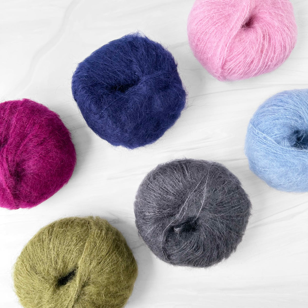 Six balls of MODE at Rowan Mohair Soft yarn in assorted colors, including pink, navy, light blue, magenta, olive green, and charcoal gray, arranged on a white wood-textured background.